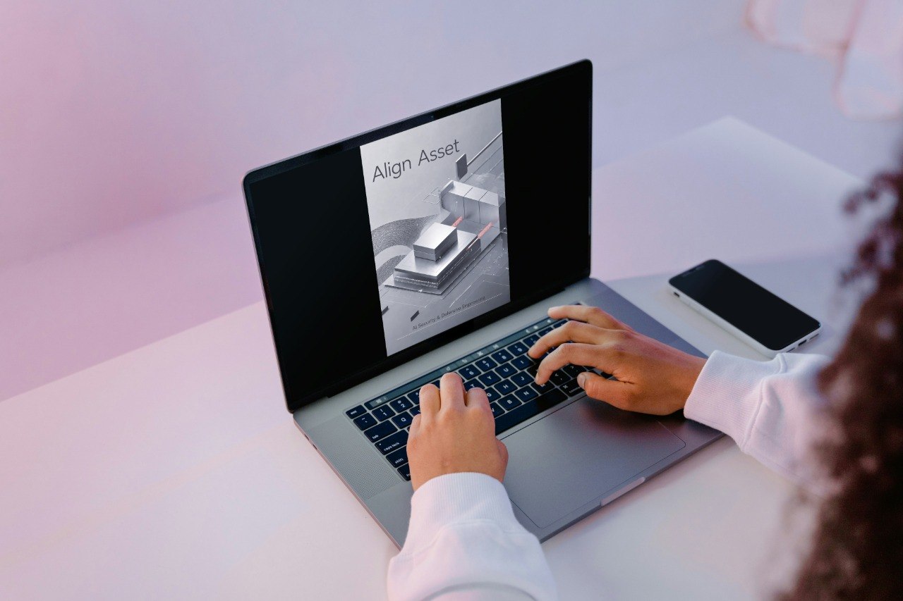 A person using a silver laptop on a light desk, with a smartphone beside it. The laptop screen displays an image labeled 'Align Asset'.