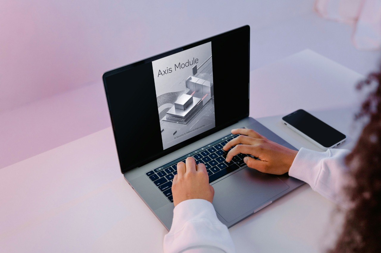 A person using a silver laptop on a white desk, with a smartphone beside it. The laptop screen displays a document titled 'Axis Module' with a grayscale technical drawing.
