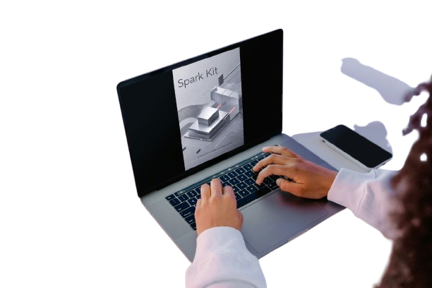 A person using a silver laptop with a black screen displaying a 'Spark Kit' manual, with a smartphone on a white surface against a white background.