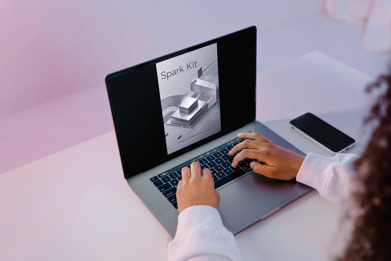 A person using a silver laptop on a light desk, with the laptop screen displaying an image for 'Spark Kit'. A black smartphone rests on the right side of the laptop, and the person's hands and white shirt sleeves are visible.